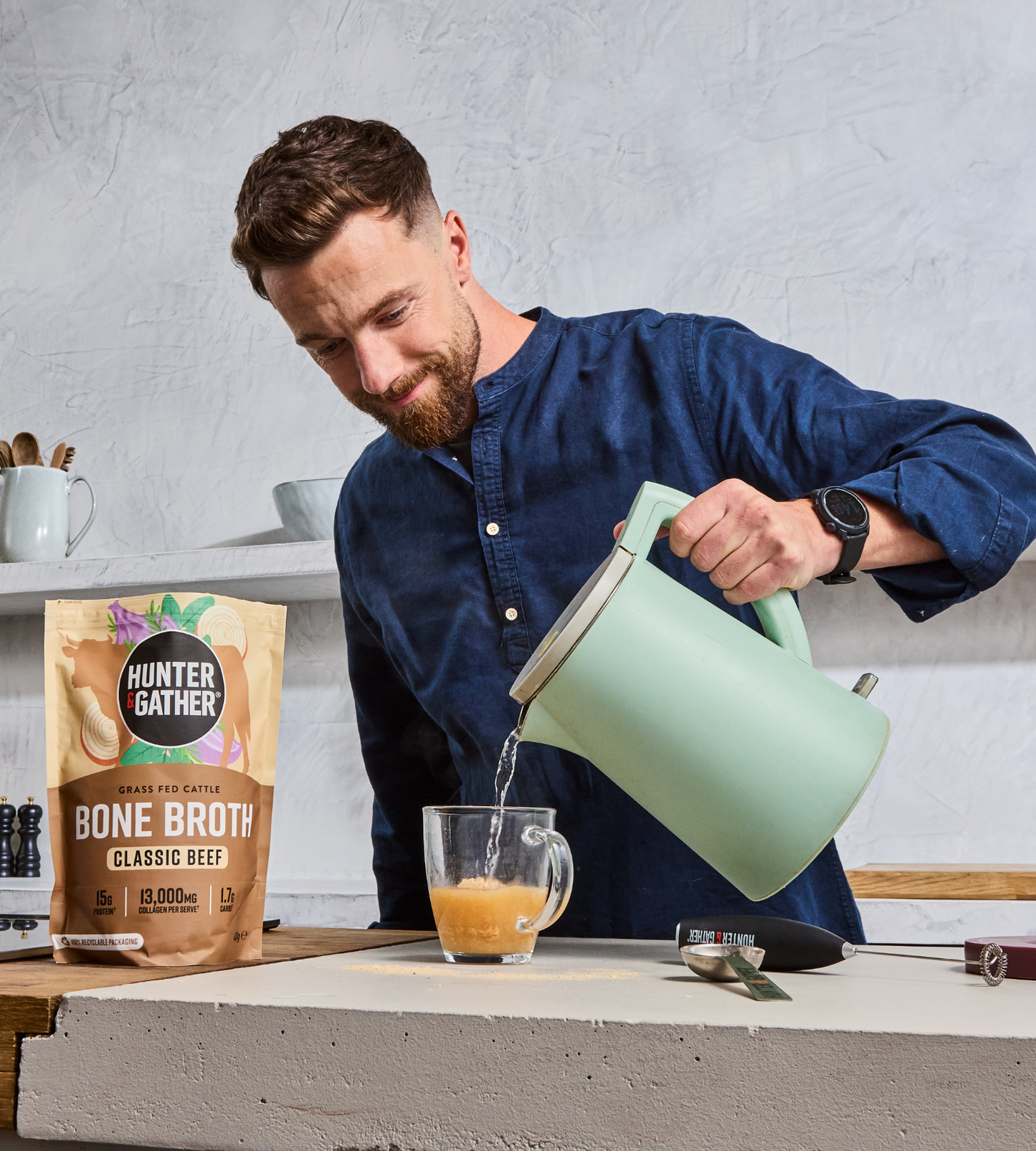 Man pouring a liquid from a green kettle into a glass, with a bag of Hunter Gather bone broth on a kitchen counter.