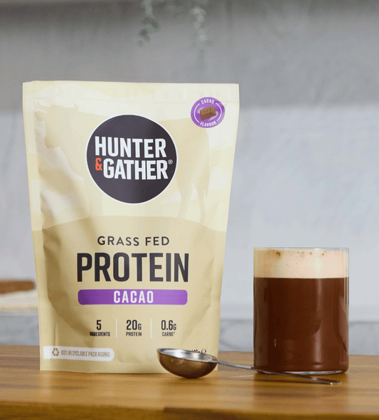 Hunter & Gather grass fed protein cacao package with a glass of chocolate shake on a wooden surface.