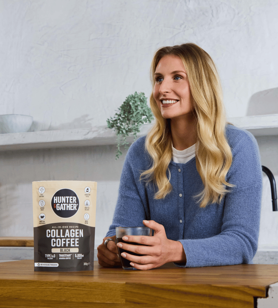 Woman holding a glass of collagen coffee with a package of Hunter and Gather Collagen Coffee on a table.