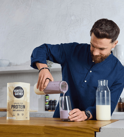 Man pouring a protein shake into a glass with a Hunter and Gather protein package on a kitchen counter.