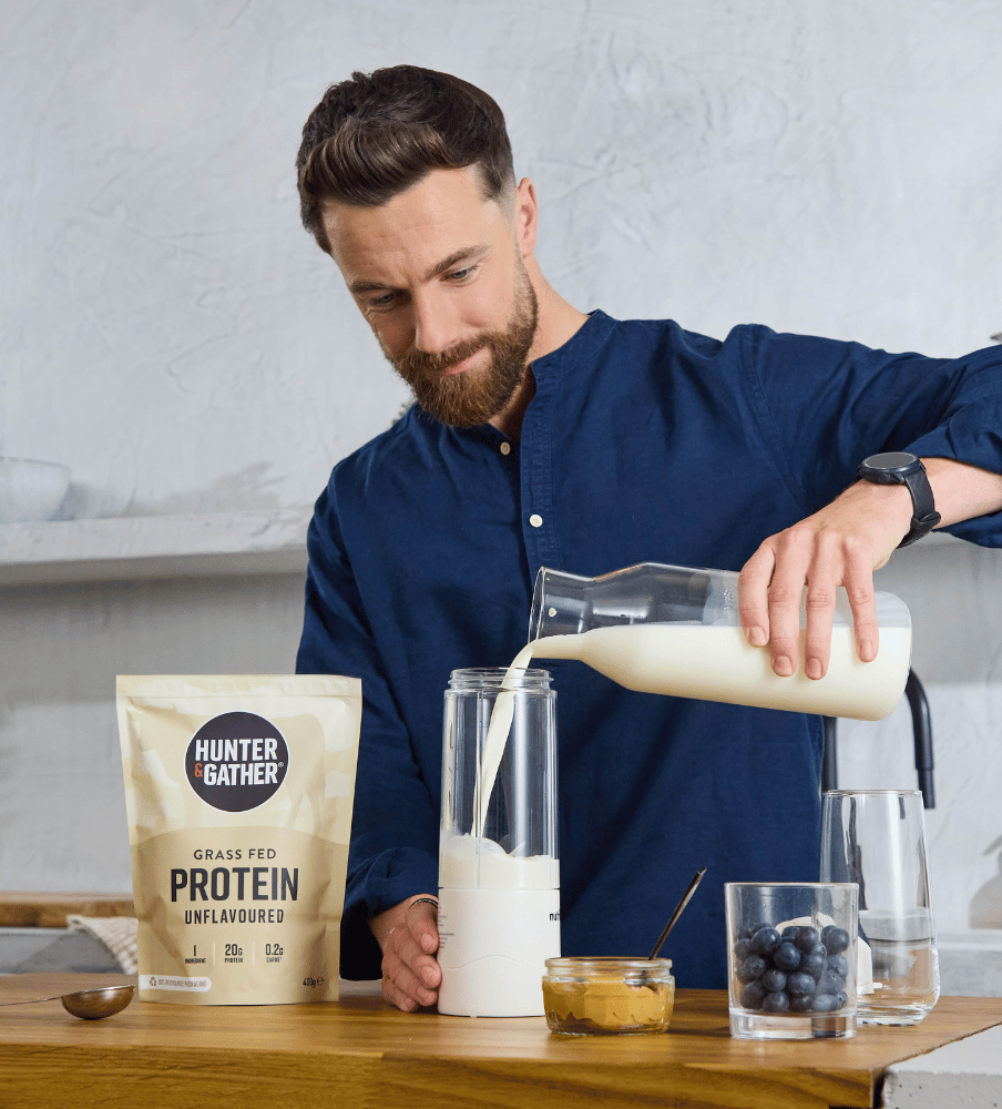 Man pouring a protein shake into a glass with a Hunter Gather protein powder bag on a wooden table.